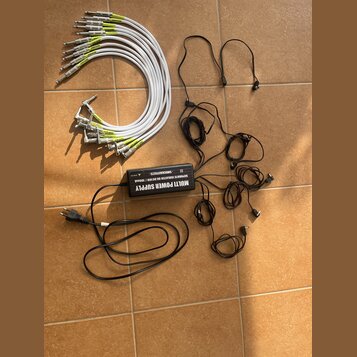 Na fotografii je černý napájecí adaptér s nápisem MULTI POWER SUPPLY, SEPARATE ISOLATED 8X DC10V / 100mA a značkou SMBCKALLIEFFECTS. K adaptéru patří…