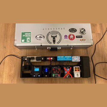 Stříbrný hliníkový kufr s různými nálepkami, včetně nápisů SPACESHIP, TONERIDER a KingTone. Pod kufrem je černý pedalboard s napájecím trafem Caline…