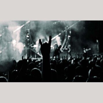 Černobílá fotografie zachycující dav lidí na koncertě. V popředí je vidět silueta natažené ruky s gestem „rocková roha“. V pozadí je osvětlené pódium…