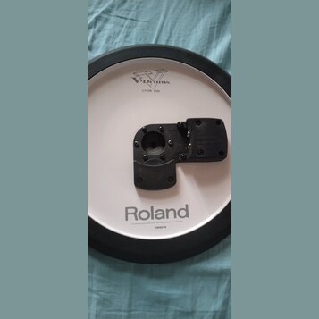Bílý elektronický činel s černým gumovým okrajem a černým plastovým úchytem v centru. Na povrchu jsou nápisy V-Drums, CY-13R Ride a Roland. Dole je u…