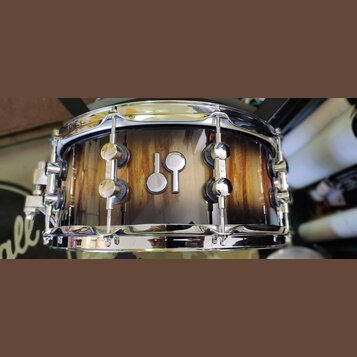 Fotografie zobrazuje snare drum s dřevěným korpusem v hnědém přirozeném odstínu s viditnou masivním kresbou dřeva. Uprostřed korpusu je umístěno stří…