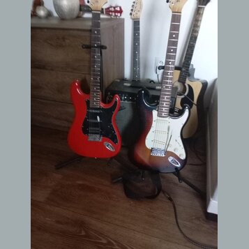 Na fotografii jsou čtyři kytary v držácích a jeden černý kombo zesilovač. V popředí jsou dvě elektrické kytary typu Stratocaster, jedna v červené bar…