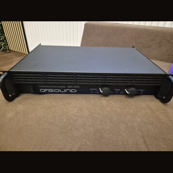 Černý výkonový zesilovač v rackovém provedení s úchyty po stranách. Na předním panelu je nápis Professional Power Amplifier QSA 600 a logo Q SOUND. Z…