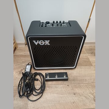 Černý kytarový kombozesilovač s bílým rámečkem kolem mřížky a logem VOX. Na horním ovládacím panelu jsou umístěny ovratidla a přepínače. Před zesilov…