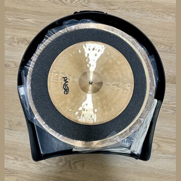 Dvě mosazné činely v černém plastovém kufru, přikryté průhlednou fólií. Na horním činele je viditelné logo Paiste. Mezi činely je umístěna černá tlum…