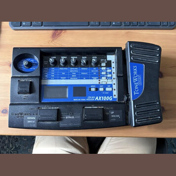 Černý kytarový procesor s modrým ovládacím panelem. Na panelu je nápis MODELING SIGNAL PROCESSOR AX100G a vpravo na boku přístroje logo TONEWORKS KOR…