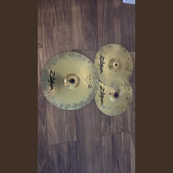 Tři mosazné činely v různých velikostech položené na hnědém laminátovém povrchu. Každý činel nese černý nápis Zildjian a logo PLANET Z. Povrchy činel…