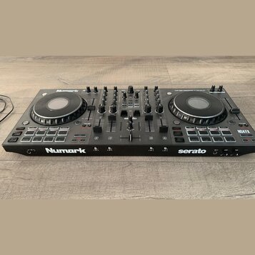Černý DJ kontrolér s dvěma jog-koly a centrálním mixážním sekcí. Na přístroji jsou viditelné nápisy Numark, serato a modelové označení NS4 FX. V horn…