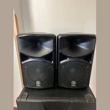 Dva černé reproduktory umístěné vedle sebe na černém transportním kufru. Oba mají v dolní části mřížku s bílým logem a nápisem YAMAHA. Povrch je v do…