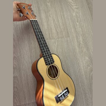 Dřevěný ukulele s přírodní barvou těla a hnědým krčkem. Na hlavě je vidět nápis CASUAL. Nástroj má čtyři struny, tmavý hmatník a černý lem kolem ozvu…