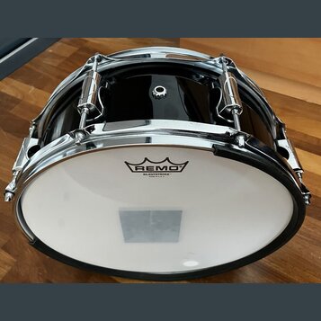 Černý buben s chromovaným obručováním a mechanismy. Na bílém koženu je vidět logo REMO a nápis SILENTSTROKE™ MADE IN U.S.A. Povrch nástroje je glossy…