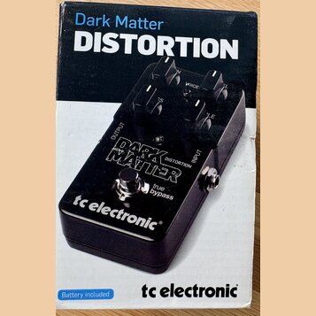 Fotografie znázorňuje krabici s efekt pedálem. Na krabici je nápis „Dark Matter DISTORTION“ a logo „tc electronic“. Zobrazený pedál je černý, má čtyř…