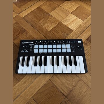 Černý kompaktní MIDI kontrolér s klávesnicí a bílými padovými tlačítky. Na zařízení jsou nápisy novation, LAUNCHKEY MINI a IN CONTROL. Povrch je v do…
