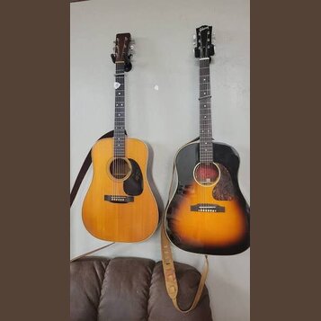 Dvě akustické kytary zavěšené na stěně. Levá kytara má světlý dřevěný povrch, černý pickguard a hnědý popruh. Pravá kytara má povrch v provedení sunb…