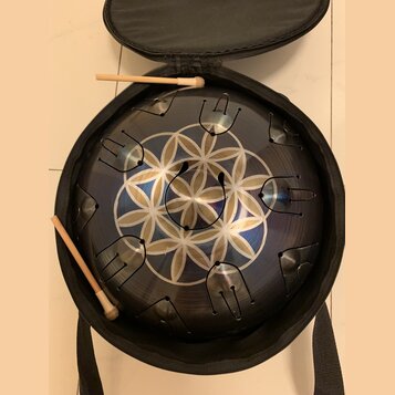 Kovový perkusní nástroj typu steel tongue drum v tmavě modré barvě s geometrickým bílým vzorem ve středu. Nástroj je umístěn v černé přepravní brašně…