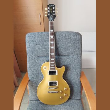 Zlatá elektrická kytara typu Les Paul s dvěma humbuckery a čtyřmi ovládacími knoflíky. Na hlavě je bílý nápis Epiphone a zlatý nápis Les Paul. Kytara…