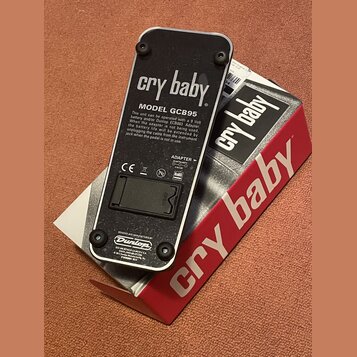 Fotografie zachycuje spodní stranu efekt pedálu v černé barvě s kovovým tělem. Na povrchu jsou nápisy „cry baby“, „MODEL GCB95“ a logo „Dunlop“. Je z…