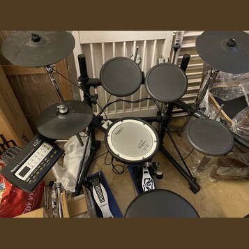 Elektronická bicí souprava značky Roland s černými pady a činely. Na module je nápis Roland TD-5 V-Drums. Centrální pad má bílý povrch s logem Roland…