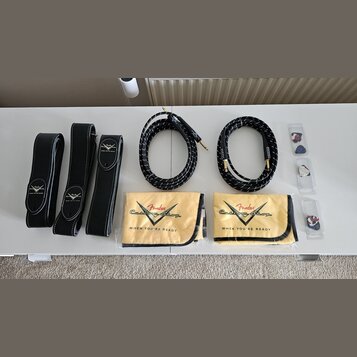 Sada černého příslušenství rozložená na bílém stole. Obsahuje tři černé kožené popruhy s bílým stehem a nápisem „Fender Custom Shop WHEN YOU'RE READY…