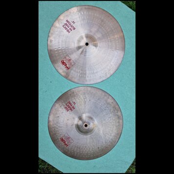 Dvě kovové činely v stříbrné barvě s viditelným broušením a otisky kladiva. Horní činel nese nápisy HEAVY HI-HAT BOTTOM 14" a logo Paiste 2002. Dolní…