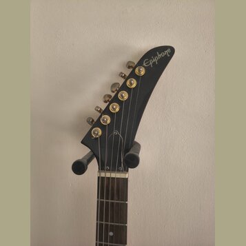 Fotografie zachycuje hlavu elektrické kytary v černé barvě. Na špičce hlavy je bílý nápis Epiphone. Nástroj má šest zlatých ladiček a hnedík z tmavéh…