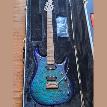 Elektrická kytara s modře gradientním tělem z plaménkového javoru, umístěná v černém tvrdém kufru. Nástroj má tři humbuckery, hned dva ovládací knofí…