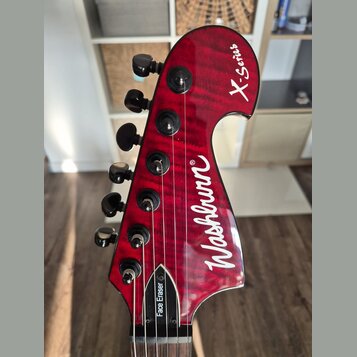 Červená hlava elektrické kytary s viditelnou strukturou dřeva. Na povrchu je bílý nápis Washburn® a v horní části označení X-Series. U dolního spoje…