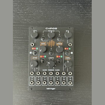 Černý modul s nápisem CHAOS v horní části a behringer ve spodní. Zařízení obsahuje několik otočných potenciometrů, tlačítka a v dolní části 12 zapušt…