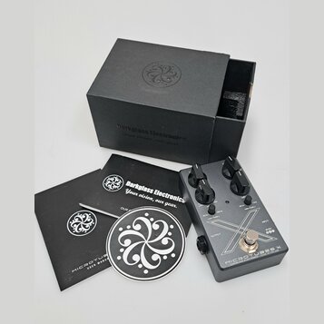 Kytarový efekt pedál stříbrné barvy s černými ovládacími knoflíky a nápisy MICROTUBES X, Darkglass Electronics - MADE IN FINLAND. Vedle pedálu je čer…