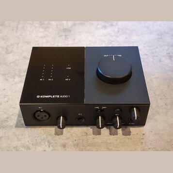 Černý zvukový interface s nápisem KOMPLETE AUDIO 1 a logem výrobce. Na horní straně jsou kontrolky s popisky IN 1, IN 2, USB a 48 V, vpravo je velký…