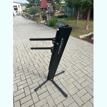 Černý kovový stojan na klávesové nástroje s dvěma vodorovnými rameny. Na hlavní nosné tyči je bílý nápis ULTIMATE SUPPORT™. Stojan stojí na dlážkovém…