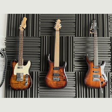 Tři elektrické kytary s hnědým plamenovitým povrchem těla zavěšené na šedém akustickém panelu. Nejlevější kytara vlevo má bílý pickguard a na hlavě l…