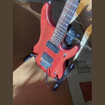 Červená elektrická kytara s tmavým hnedým hmatníkem, umístěná na černém stojanu. Na kytaru jsou instalovány dva humbuckery, přičemž na horním snímači…