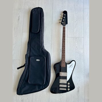 Černá elektrická basová kytara s bílým plechtrátorem a hnědým hmatníkem. Na hlavě nástroje je svisle napsáno logo "Rickenbacker". Vedle kytary leží č…