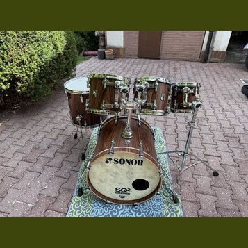 Bubnová sada v tmavě hnědé dřevěné povrchové úpravě. Na basovém bubnu je bílá kožená blana s nápisy SONOR a SQ2 DRUM SYSTEM. Na ostatních bubnech jso…