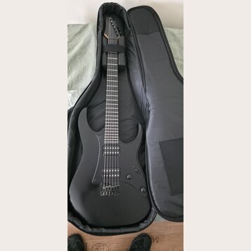 Černá elektrická kytara s matným povrchem v černém softcasu. Nástroj má dva humbuckery, fixační most a na hlavě není vidět žádné logo ani nápis. Povr…