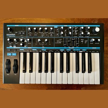 Černý analogový syntetizátor s klaviaturou a ovládacím panelem. Na levé straně je nápis novation BASS STATION II Analog Synthesizer. Panel obsahuje m…