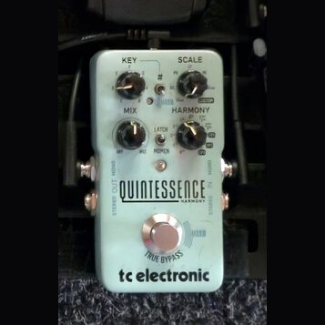 Světle modrý efekt pedál s nápisy QUINTESSENCE HARMONY a tc electronic. Na horní části jsou tři černé otočné ovladače s popisky KEY, SCALE a MIX, dál…