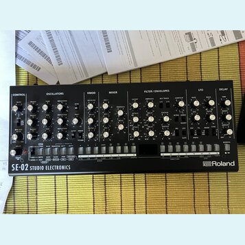 Černý analogový syntetizátor s nápisem SE-02 STUDIO ELECTRONICS a logem Roland. Přední panel obsahuje numeroustočítka, přepínače a sekci s klávesovým…