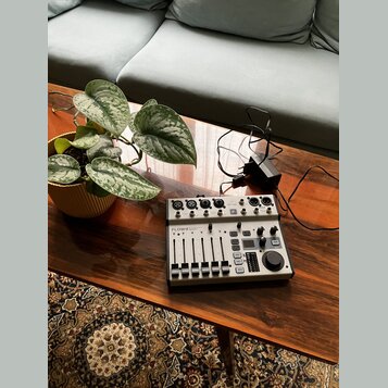 Na dřevěném stole je umístěn kompaktní mixážní pult stříbrné barvy s černými ovládacími prvky. Na zařízení je viditelné logo Behringer a nápis FLOW 8…