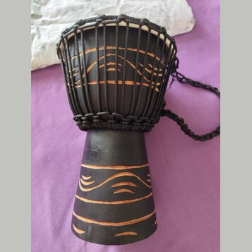 Černý djembe s bílou membránou, nataženou pomocí černých provázků. Tělo nástroje je zdobeno vyrytými vodorovnými a vlnitými liniemi, které odhalují h…