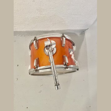 Oranžový buben s kovovými obručemi a lugy. Na korpusu je vidět logo s nápisem DUGGERY, které je v pravé části částečně odloupané a poškozené. K bubnu…