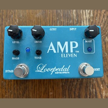 Modrý kytarový efekt s bílými nápisy AMP™ ELEVEN a Lovepedal CUSTOM EFFECTS. Na horní straně jsou tři černé otočné knoflíky s popisky LEVEL, DRIVE a…