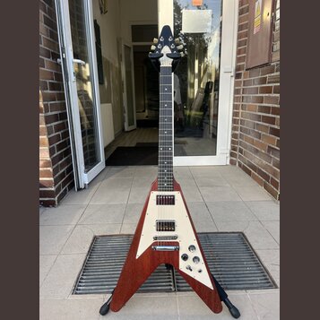 Elektrická kytara s tělem ve tvaru V v červenohnatém dřevěném provedení s bílým plechem. Nástroj má dva humbuckery, tři otočné knofíky a bílé ladicí…