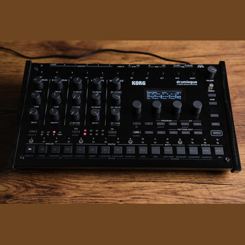 Černý hybridní bicí automat KORG drumlogue. Zařízení obsahuje množství otočných knoflíků, několik tlačítek a malý displej s textem BASS DRUM. Na horn…