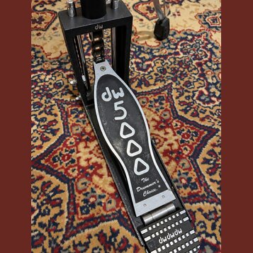 Černý bicí pedál s kovovými prvky. Na šlapadle je bílým nápisem značka dw a model 5000, dále text The Drummer's Choice.® a logo dw na horní části kon…