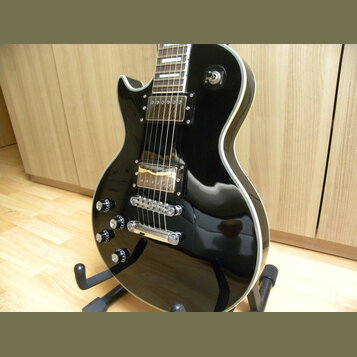Černá elektrická kytara s humbuckery a bílým lemováním kolem korpusu. Na kytaru jsou vidět čtyři ovládací knofíky s nápisy „VOLUME“ a „TONE“. Nástroj…