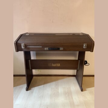 Hnědý klavír nebo digitální piano v dřevěném korpusu s uzavíracím víkem. Na přední spodní části je zlatý nápis „FAKFISA“ s doprovodným logem. Nástroj…