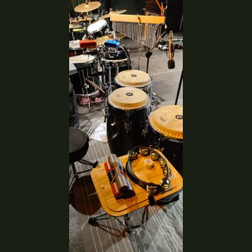 Fotografie zachycuje soubor perkusních nástrojů a bicí sadu. V popředí je dřevěný stolek, na kterém leží tamburína s kovovými cinkety a dva šedé s če…