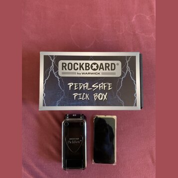 Na fotografii je krabička a dvě součásti příslušenství. Na krabici je nápis ROCKBOARD® by WARWICK a ručně napsaný text PEDALSAFE PICK BOX. Vedle krab…